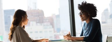 Two women having a meeting. They are sitting at a table across from each other with highrise buildings visible in the background.