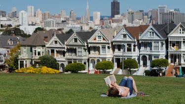 A woman reads a book on a grassy area. The city of San Francisco can be seen in the background.