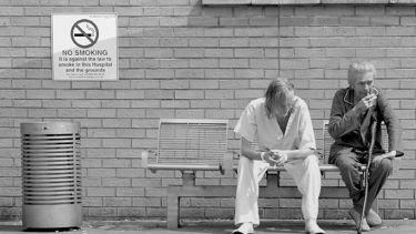 Two people sit and smoke next to a no smoking sign outside a hospital.