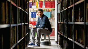 An image of a reader sat on a kickstool looking at shelves of books.