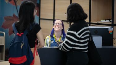 Two people standing at talking to the third person sitting at the Library Welcome Desk 