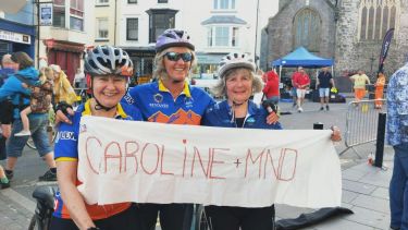 Sara, Glynis and friend on their bicycles holding a banner reading: Caroline + MND