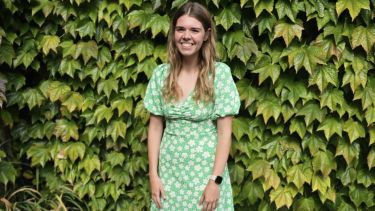 Charley-Ann stood in front of a hedge smiling in a green floral summer dress