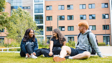 Three students sat on the grass, there is one of the student accommodation buildings in the background