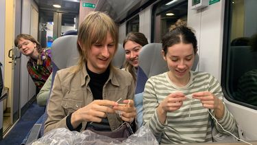 on a train: two students in the foreground crocheting and 2 other looking form behjind the second row