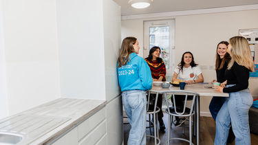 A group of girls stood around a table in a flat kitchen, one is in a blue hoodie with Mentor 2023-24 written on the back