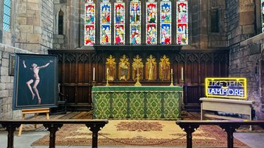 Photo of the high altar at Rotherham Minster,. The altar has a green frontal and stands in front of reredos, above which are a series of stained glass windows. To the left of the altar is a painting on an easel depicting a nude, white man against a dark background, suspended by a web of thorns. To the right of the altar is a yellow and lilac neon text piece which reads 'Like this I am more' across two lines.