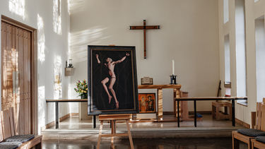 Photo of a chapel with an altar in the background and a painting in the foreground, left of centre. The painting is on an easel and depicts a nude, white man against a dark background, suspended by a web of thorns. Windows on the right hand side cast light on the wall and door on the left hand side of the image.