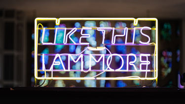 Close up photo of a yellow and lilac text piece which reads 'Like this I am more' across two lines. Stained glass windows are visible through the spaces between letters.