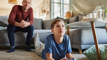A boy holds a video game controller and is looking off camera to where he is playing a video game. His father sits behind him on a sofa watching him play.
