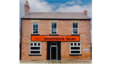 Red brick two storey house with chimney 3 upstairs windows and 2 downstairs windows either side of a black door.  A large rectangular orange Henderson's relish sign sits between the windows of the ground and first floors.