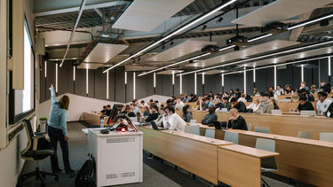 A person stands with their back to the camera, as they are pointing to a large screen/projector in front of a lecture theatre of seated students