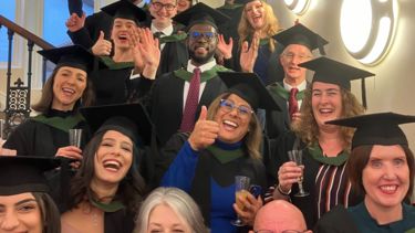 A group of graduates stand on stairs in their gowns and wave at the camera