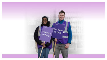 Two students in hi-vis jackets holding signs reading I'm here to help