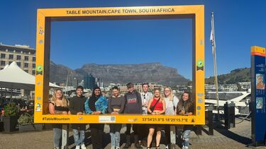 A group of students smiling at the camera whilst stood behind a Table Mountain, Cape Town, sign