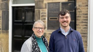 A woman with short grey hair wearing dark framed glasses and a flowing grey cardigan and a man with brown curly hair stand smiling outside the entrance to a stone building.