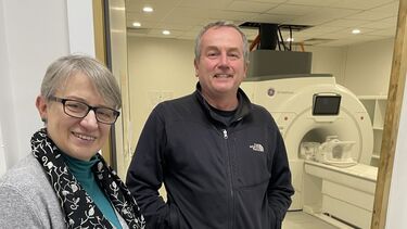 A woman with short grey hair wearing dark framed glasses and a flowing grey cardigan and a man with short grey hair wearing a black top stand smiling in front of an MRI scannner