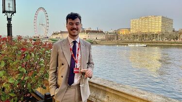 A man with brown curly hair wearing a beige suite and a red visitor pass stands holding a glass of wine in front of the river thames with buildings and the london eye observation wheel in the background.