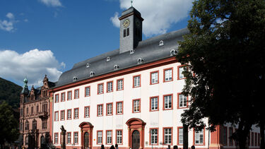 Large, old building with clocktower is lit up by sunshine. People walk across the courtyard infront