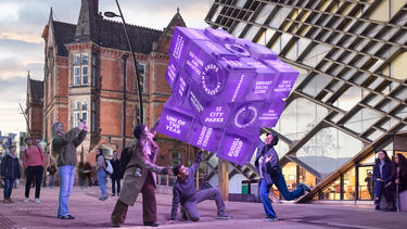 Four students interact with a large floating Rubik's cube outside two University buildings (The Diamond and Jessop Building)