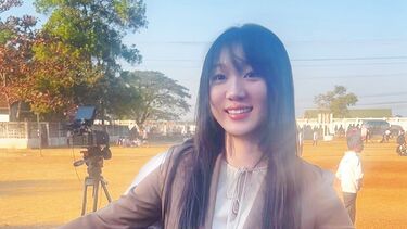 A young woman stands smiling at the camera in an outdoor space whilst a group of young children gather round her to hug her.