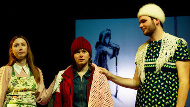 three students on stage, the outside 'grannies' lie their hands on the shoulders of the middle one