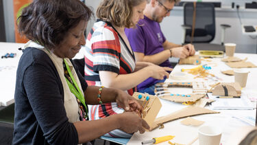 Three people sit creating musicall instruments out of cardboard and craft materials