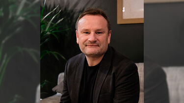 A headshot of Jez Groom smiling on a sofa with a houseplant in the background