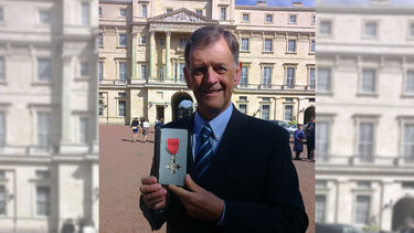 Mel Evans in a blue suit, standing in front of Buckingham Palace holding his MBE medal