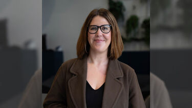 A headshot of Sarah Flude in an office, smiling in a brown jacket