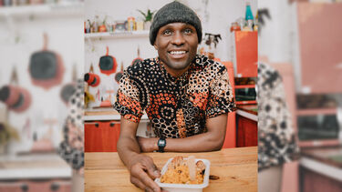 Tendai Taruvinga sitting at a kitchen counter with a tray of food, wearing a bright patterned shirt and a beanie hat
