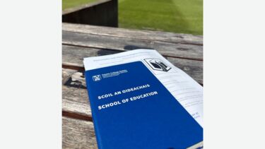 A book with a sheet of paper tucked into it. It reads "Scoil an Oideachais - School of Education'