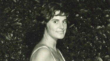 Black-and-white headshot of a young Maureen Halton smiling, wearing a white dress with her dark hair in a short bob, and standing against a leafy hedge