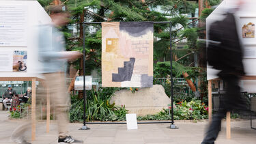 A blurry, long-exposure image of two people walking past a textile art piece displayed on a stand. The textile piece features abstract geometric shapes in various colors and textures, and it's positioned between two white display boards with text.