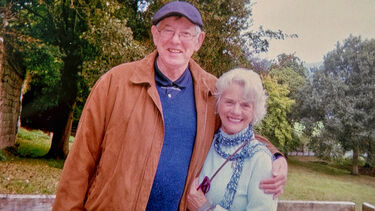 Christopher Ball with his arm around Gail Ball on a wooden veranda in a tree-lined park