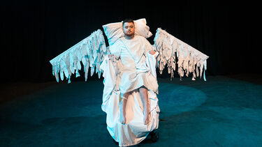 Jamie Hale is on a stage in their wheelchair. They are dressed in white hospital gown and are wearing a large pair of white feathery wings.