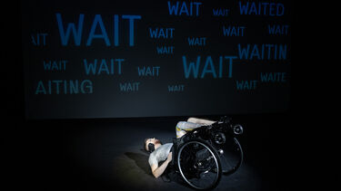 Jamie Hale is on a stage laid on their back in their wheelchair. The background is black with illuminated in blue the word wait multiple times.
