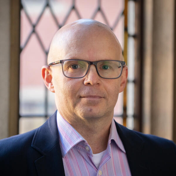 Profile picture of A person without hair wears glasses. They're in front of leaded windows