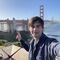 A male student stands outside with the Golden Gate Bridge in the background. He is smiling at the camera and making a peace sign with his hand.