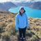 A female student wearing a blue winter hat and blue waterproof jacket stands smiling at the camera on a hill. In the background is a large lake with mountainous backdrop.