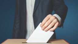 A white person wearing a suit, putting a ballot paper into a slot in a wooden box