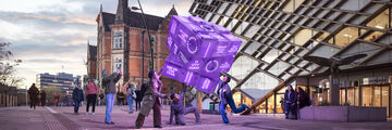Four students interact with a large floating Rubik's cube outside two University buildings (The Diamond and Jessop Building)