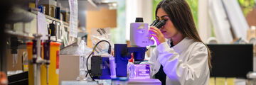 A University of Sheffield student looking through a microscope at a glowing cube that has research-led teaching written on it. 