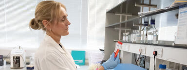A woman in a research lab, wearing a lab coat, holding a tube of liquid.