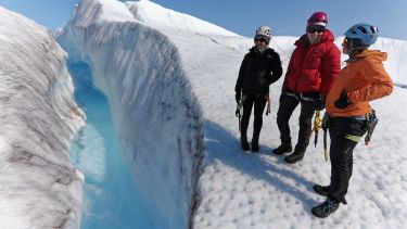 Three researchers stand on a glacier looking into a ravine