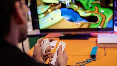 A photograph shows adult hands holding a set of Nintendo Wii controllers and playing a video game on screen.
