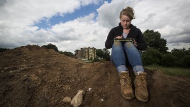 A student wearing outerwear and hiking boots sits by an excavation site using a tablet.