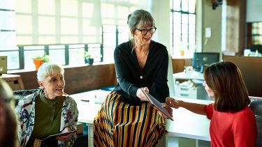 Three women round a desk two are sharing information via a tablet. Two of the women are older