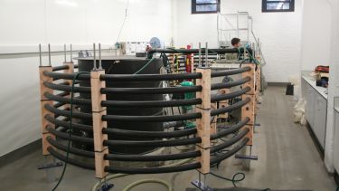 A row of black pipes create a curved frame around technical equipment inside a bright research facility