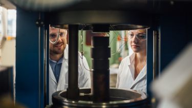 Two engineers in lab coats seen through a glass-walled machine with tangles of wires sticking out